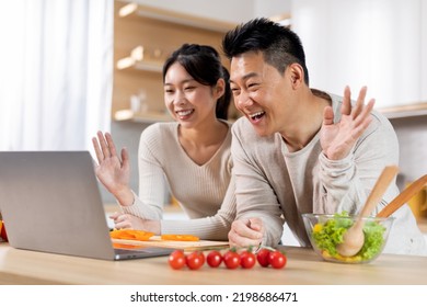 Happy Asian Couple Having Video Chat With Friends Or Family While Cooking Together At Kitchen, Standing Next To Table Full Of Vegetables, Waving And Smiling At Computer Screen, Copy Space