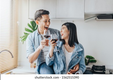 Happy Asian Couple In Love Laughing And Drinking Red Wine In The Kitchen To Celebrate Their Anniversary