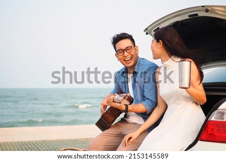 Happy Asian couple lover having stop off by the sea and sitting in hatchback  ,they are  drinking coffee playing guitar together.