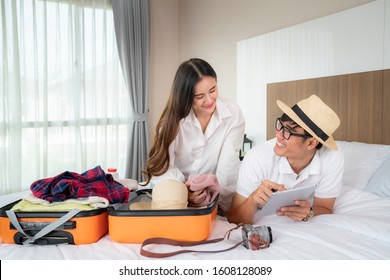 Happy Asian Couple Packing Suitcase On The Bed In Room Use Tablet For Search Travel Trip Online

