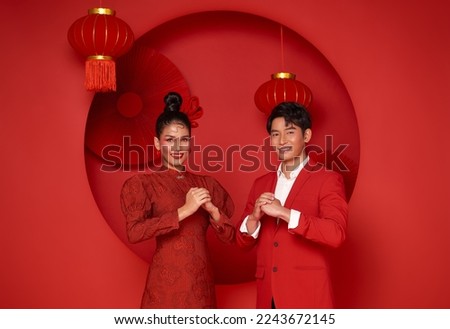 Happy asian couple in red casual attire with gesture of congratulation greeting happy new year 2023 on bright red background.