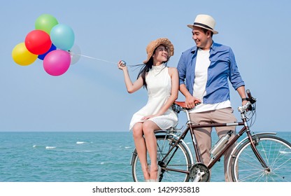 Happy Asian Couple Riding Bicycle And Holding Colorful Balloons On The Beach, Lifestyle Concept.