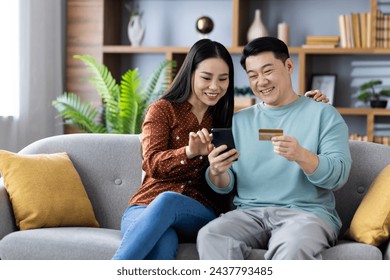 Happy Asian couple sitting on the couch with a smartphone and credit card, smiling and enjoying online shopping in a cozy home setting. - Powered by Shutterstock