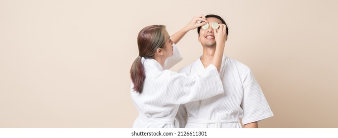 Happy Asian couple smile in bathrobe or spa suit doing cucumber slice facial mask together on banner brown isolated studio background.