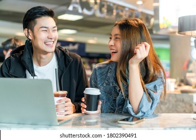 Happy Asian Couple Take Off Face Mask In A Coffee Shop Surfing Internet On Laptop. Young Man And Woman In A Restaurant Looking At Touch Screen Computer Laugh Smile Together