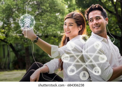 Happy Asian Couple Using And Listening The Song And Using The Social Network Via Smart Mobile Phone For Online Shopping With Omni Channel In The Park, Lifestyle And Technology Concept