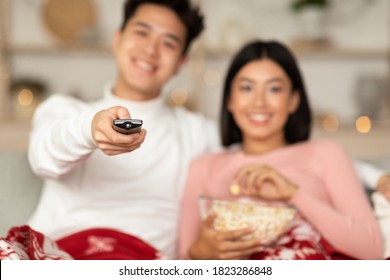 Happy Asian Couple Watching TV On Christmas, Enjoying Favorite Xmas Movies And Popcorn Sitting On Couch At Home, Focus On Remote Control. Family New Year Eve Leisure. Shallow Depth