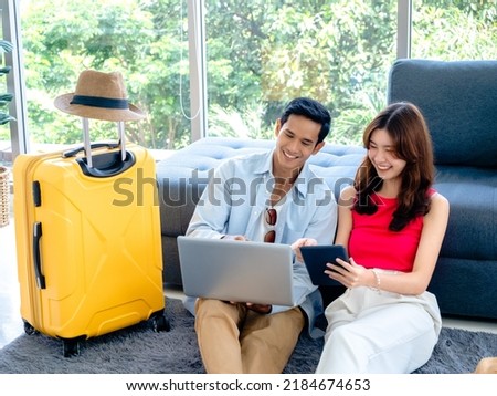 Happy Asian couple, young man and woman looking and using laptop computer and smartphone together for flight booking and trip information in living room near luggage, travel holiday summer vacation.