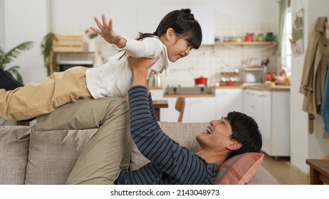 Happy Asian Dad Having Fun Lifting Up Daughter On Straight Legs While Playing Airplane On Living Room Couch At Home. Father Doing Yoga With Child Concept