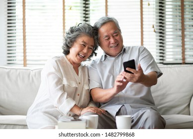 Happy Asian Elderly Couple Laughing Sitting On White Couch While Using Smarting Technology At House. Senior Lover Enjoying While Doing Video Call Via App On Phone. Older Japanese People Having Fun