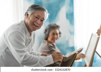 Happy Asian Elderly Couple Painting On Canvas At Their House On Holidays. Artist Senior Man And Woman In Casual Looking At Camera. Retirement.