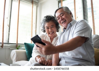 Happy Asian Elderly Couple Sitting On White Couch And Laughing While Using Smarting Technology At Their House. Senior People Enjoying While Doing Video Call Via App On Phone.