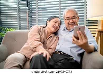 Happy Asian Elderly Couple Sitting On Couch While Using Smartphone Video Call Online Together In Living Room, Holiday Activities For Retired Couples, Senior Health Care And Wellness Mental Concept.