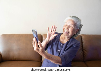 Happy Asian Elderly Woman With Smartphone Having Video Call And Waving Hand In Living Room At Home. Technology And Older People Concept.