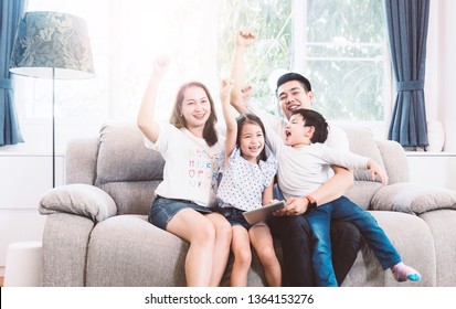 A Happy Asian Family Cheer Up And Do Activity Together In Living Room Sitting On Sofa At Home. Father Is Holding A Tablet Play Game With Daughter And Son.