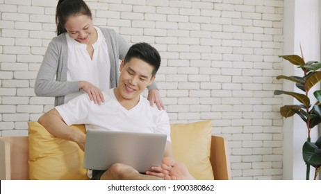 Happy Asian Family Couple Husband And Wife Laughing Sitting On Sofa Using Laptop Computer Working From Home Business Man Sitting On Sofa Home Working On Laptop Her Woman Is Giving Her Shoulder Massage