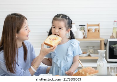 Happy Asian family, cute little daughter eating bread with jam, rainbow sugar flakes feeding from young mother after spreading jam on sliced bread together, preparing breakfast in kitchen at home - Shutterstock ID 2172808063