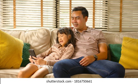 Happy Asian Family Dad And Little Kid Daughter Using A Smart Phone And Having A Video Call Chatting Or Taking A Selfie Sitting On A Couch Fun And Enjoying Online Communication In Living Room At Home.