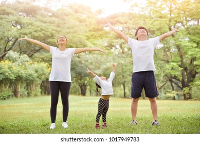 Happy Asian Family With Daughter In White Clothes Putting Their Hands In The Air While Warm Up At Park. Young Man, Woman And Little Kid Girl With Open Arms Gesture Exercising At Outside. Having Fun