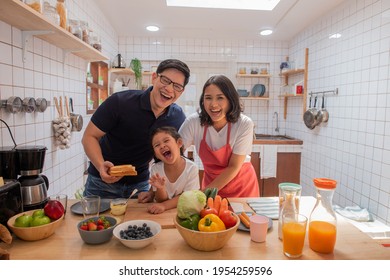 Happy Asian Family Eat Breakfast Together In Kitchen. Asian Couple With Cute Baby Daughter Preparing Food For Breakfast Meal. Mom And Dad With Child Girl Kid Enjoy And Having Fun Cooking Together.