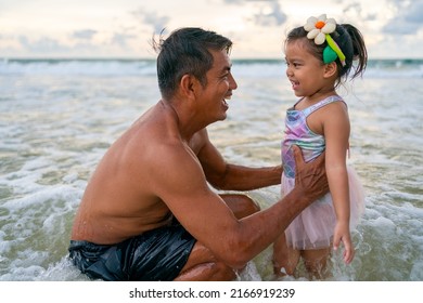 Happy Asian Family Grandfather Carrying And Playing With Little Grandchild Girl While Walking On The Beach On Summer Vacation. Senior Man And Child Girl Kid Enjoy Outdoor Lifestyle Together At The Sea