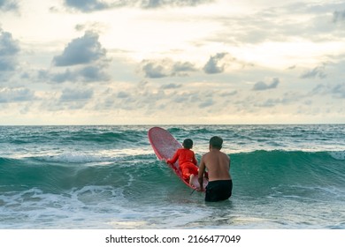 Happy Asian Family Grandfather Teaching Grandchild Boy Surfing On Surfboard In The Sea Summer Vacation. Senior Man And Little Boy Enjoy Outdoor Activity Lifestyle And Water Sport Surfing Together.