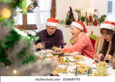 Happy Asian Family, Grandparents And Mother With Santa Claus Hats Eating Christmas Dinner, Talking And Smiling, Having Good Time Together In A Christmas Decorated Dining Room At Home.