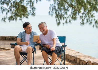 Happy Asian Family Handsome Man Teach Senior Father Using Digital Tablet For Video Call Or Online Shopping On The Beach In Summer Day. Father And Son Enjoy And Having Fun Together In Holiday Vacation