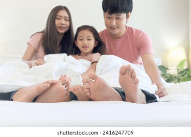 Happy Asian family has fun in bedroom. Feet of mother, father and daughter lying in bed together and hiding under white blanket. Parents and kid girl have good memory at home. Parenthood and childhood - Powered by Shutterstock