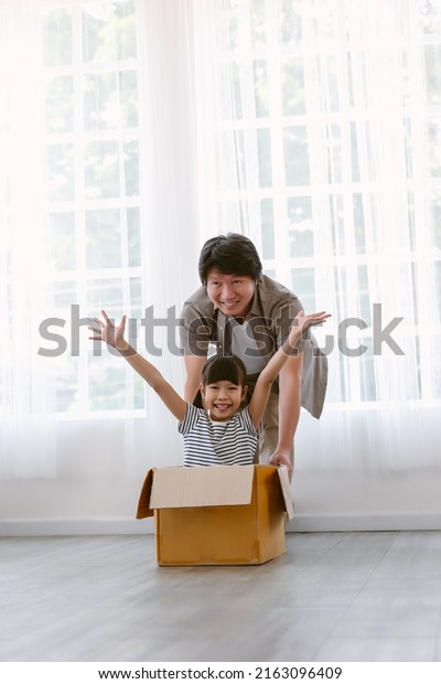 Happy Asian family have a fun spend time at\
home. Portrait of father and adorable daughter pushing cardboard\
box around living room in junk modelled car. Real estate, purchase,\
Family concept.