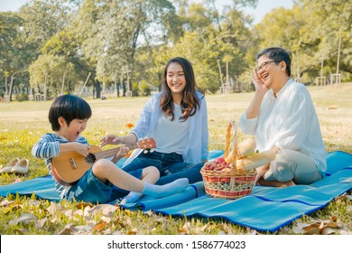 Happy Asian Family Having Fun Picnic In Park Autumn Together. Mother Touching To Kid With Smile While Grandmother Looking At Child Playing Ukelele With Happiness. Relax Family Activity.