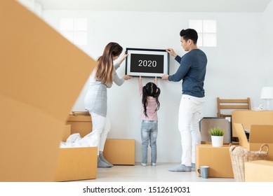 Happy Asian Family Holding And Putting Picture Frame On The Wall. Young Man, Woman And Little Kid Girl Decorating Room Together. 2020 To 2021, New Year Concept. Moving House.