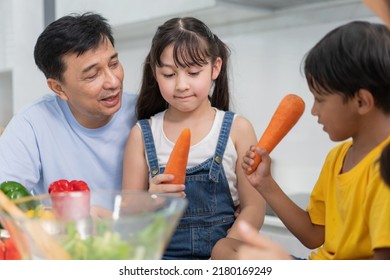 Happy Asian Family Making Salad In The Kitchen Together. Father Supporting Kids To Learn Cooking. Playful Son Playing With Vegetables With His Sister. Activity On Weekend