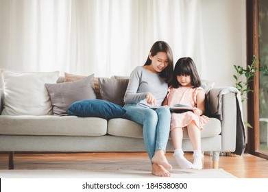 Happy Asian family mother and daughter sitting on sofa reading a book in living room at home - Powered by Shutterstock