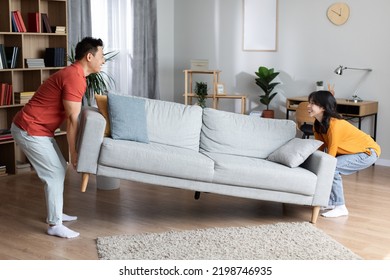 Happy Asian Family Moving Furniture At Their New House, Positive Middle Aged Husband And Young Wife Pushing Sofa In Cozy Living Room, Enjoying New Apartment, Full Length, Copy Space