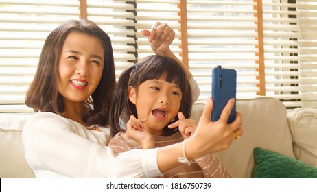 Happy Asian Family Mum And Little Kid Daughter Using A Smart Phone And Having A Video Call Chatting Or Taking A Selfie Sitting On A Couch Fun And Enjoying Online Communication In Living Room At Home.