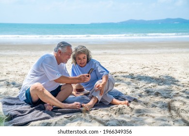 Happy Asian family on beach vacation. Senior couple sit on the beach using smartphone for online shopping or video call. Healthy elderly husband and wife enjoy outdoor lifestyle together in summer. - Powered by Shutterstock