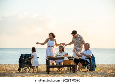 Happy Asian Family On Holiday Vacation. Group Of Multi Generation Family Little Girl With Parents And Grandparents Relax And Enjoy With Dinner Party And Dancing Together On The Beach At Summer Sunset.