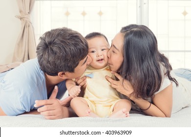 Happy Asian Family Parents Kissing Their Baby Boy In Bedroom