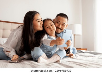 happy asian family with phones sit at home on the bed and communicate via video call, little korean girl with mom and dad takes selfie on smartphone, daughter takes picture of herself with her parents - Powered by Shutterstock