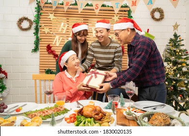 Happy Asian Family With Santa Claus Hats, A Middle-aged Son Along With His Wife And Father Giving X'mas Present Box To Mother During Christmas Dinner Celebration In A Christmas Decorated Dining Room.