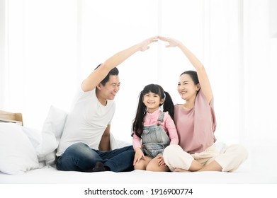 Happy Asian Family Sitting On Bed In Bedroom Together And Making The Home Sign. Mom And Dad Making Roof Figure With Hands Arms Over Heads