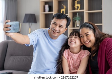 Happy Asian Family With Teen Child Daughter Having Fun Taking Selfie On Mobile Smartphone At Home. Smiling Parents And Teenage Kid Girl Embrancing And Posing For Take Family Photo On Sofa.