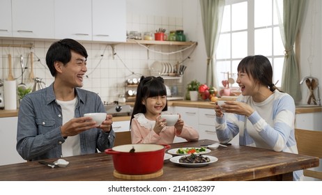 Happy Asian Family Of Three Enjoying Hot Soup From Bowls In A Modern Dining Room With Daylight At Home. Everyone Laughs While The Mother Says Yummy