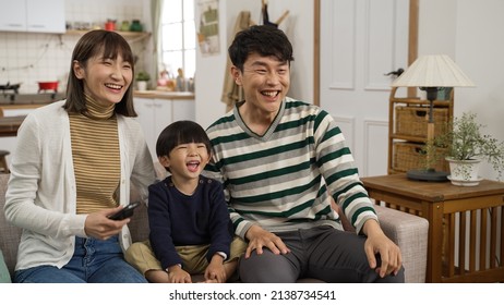 Happy Asian Family Of Three Having Fun Watching Television Together At Home. Cute Boy Laughing With His Father And Mother While They Are Pointing At The Tv