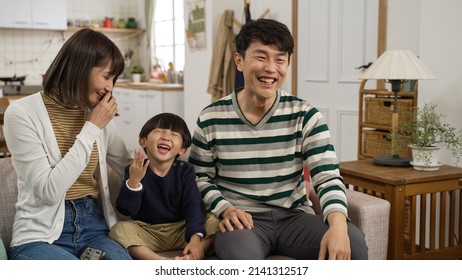 Happy Asian Family Of Three Having Fun Watching Television Together At Home. Cute Boy Laughing With His Father And Mother While They Are Pointing At The Tv