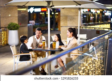 Happy Asian Family With Two Children Relaxing Talking Having Drinks And Desserts At An Outdoor Coffee Place.