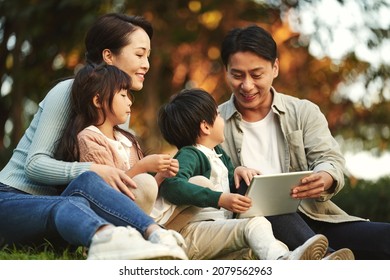 Happy Asian Family With Two Children Having A Good Time Sitting On Grass Relaxing In City Park At Sunset