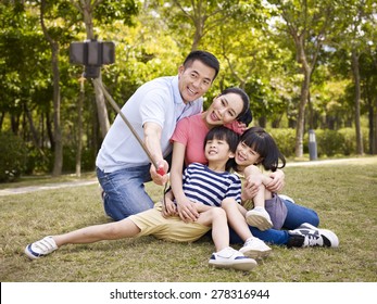 Happy Asian Family With Two Children Taking A Outdoor Selfie With Selfie Stick Outdoors In A City Park.