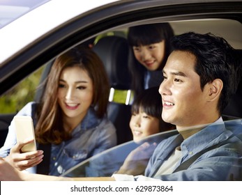 Happy Asian Family With Two Children Traveling By Car, Mother Using Cellphone While Father Driving, Focus On The Father.